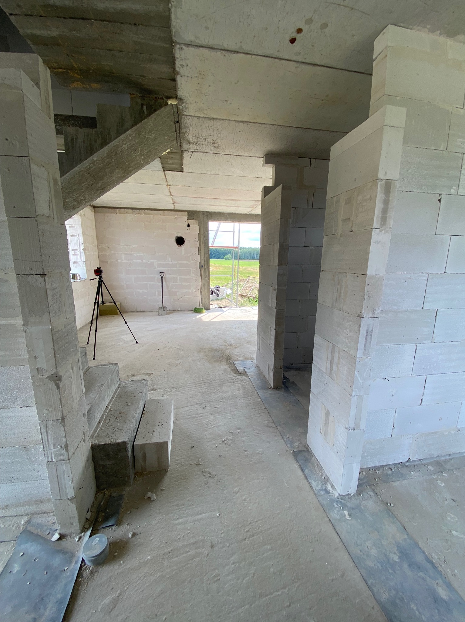 Wnętrze w trakcie budowy: ściany z białych bloczków, betonowy strop, drewniana belka wzmacniająca. W tle widoczny krajobraz przez otwór okienny.