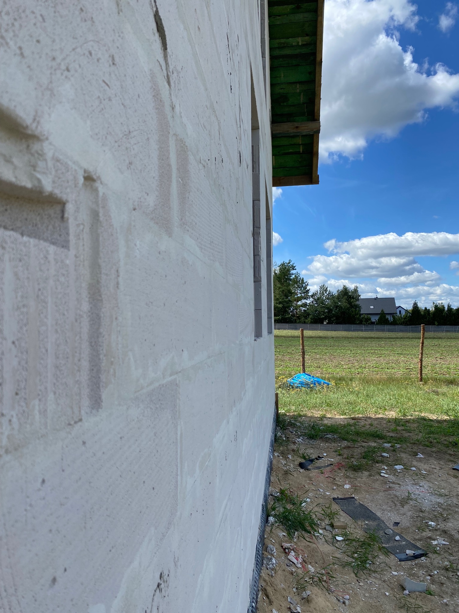 Ściana budynku w trakcie budowy, wykonana z białych bloczków, widok z boku. W tle zielone pola i niebo z chmurami. Widoczne drewniane elementy konstrukcji dachu.
