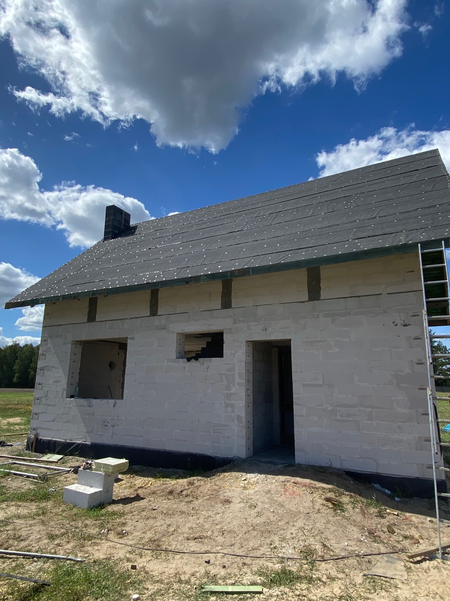 Stan surowy domu z białych pustaków w Lucynowie Dużym. Widoczne okna, drzwi i czarny dach z papy. Na ziemi leżą materiały budowlane, a obok budynku stoi drabina.