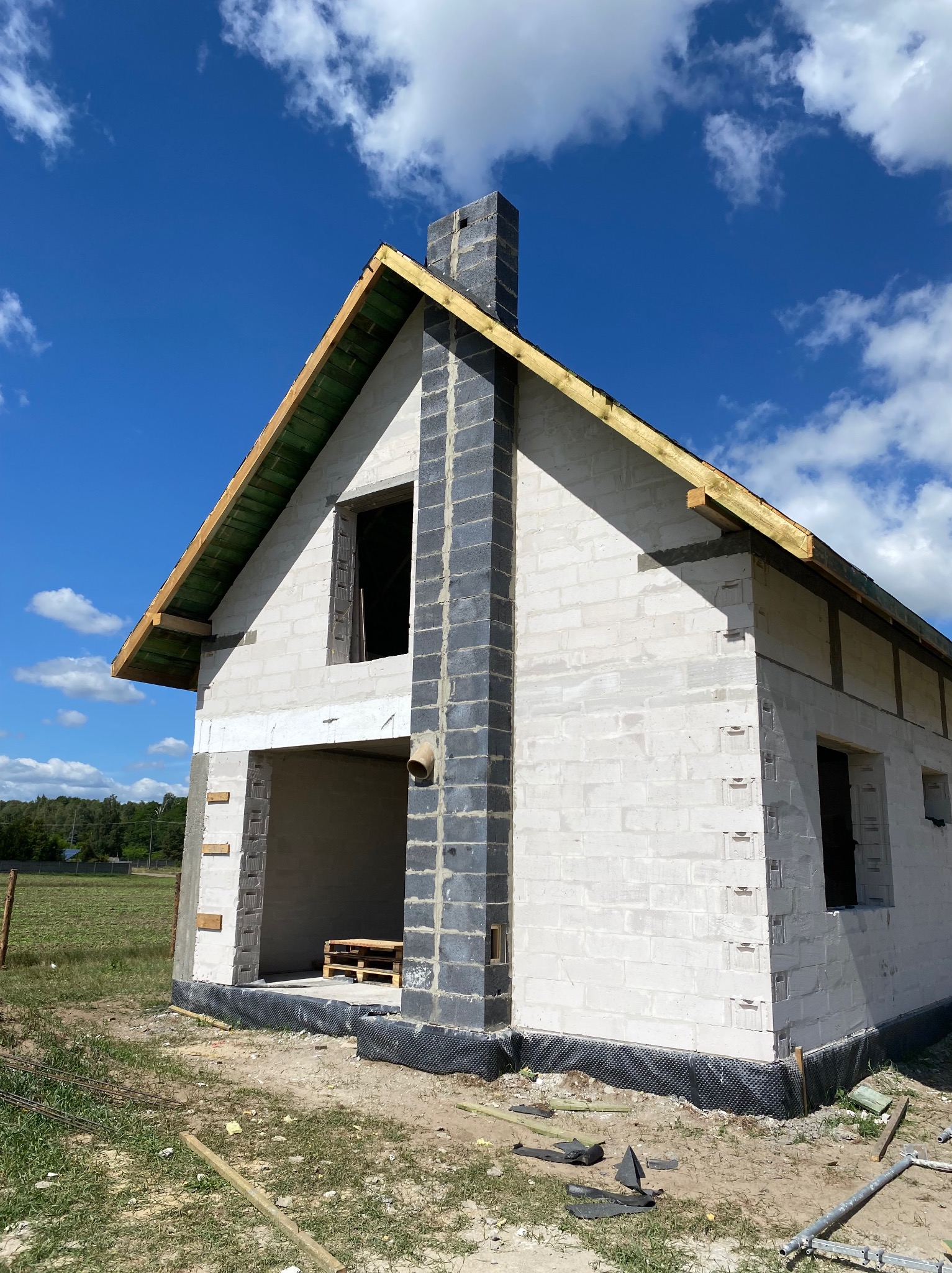 Niewykończony dom z białych bloczków z ciemnym kominem, widok z zewnątrz w słoneczny dzień. Widoczne palety w otworze drzwiowym i elementy konstrukcji dachu.