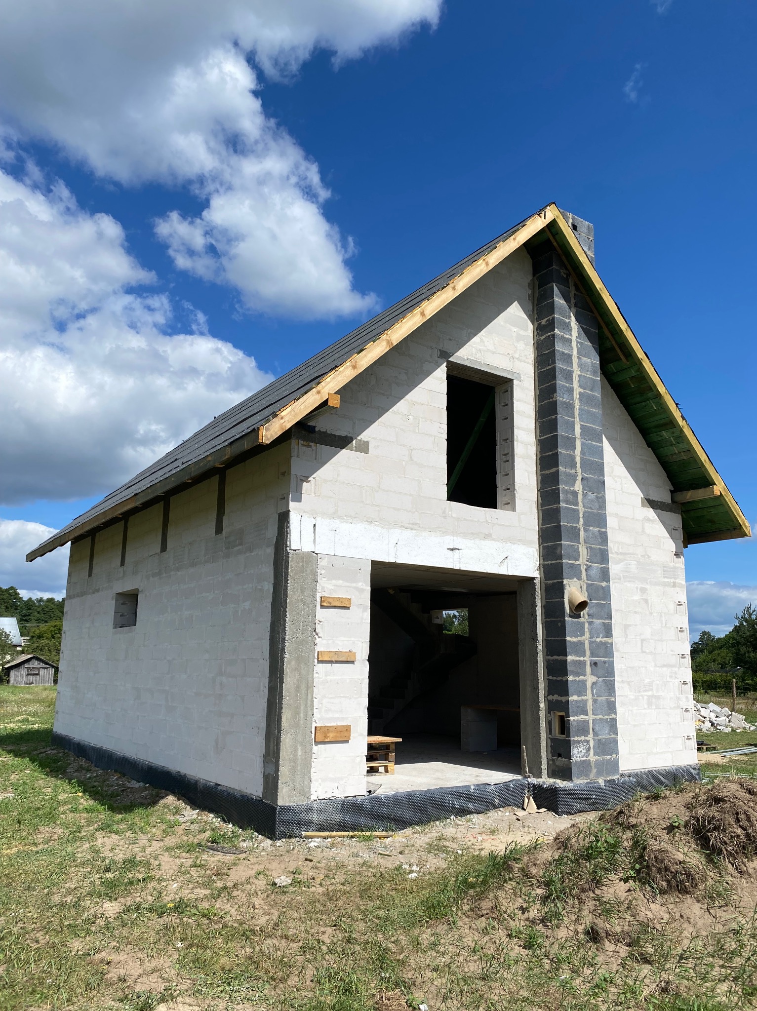 Surowy stan domu z białych bloczków, z widocznym kominem z szarych bloczków. Dach pokryty papą, widoczne drewniane elementy konstrukcyjne. Okno i otwór drzwiowy bez stolarki.