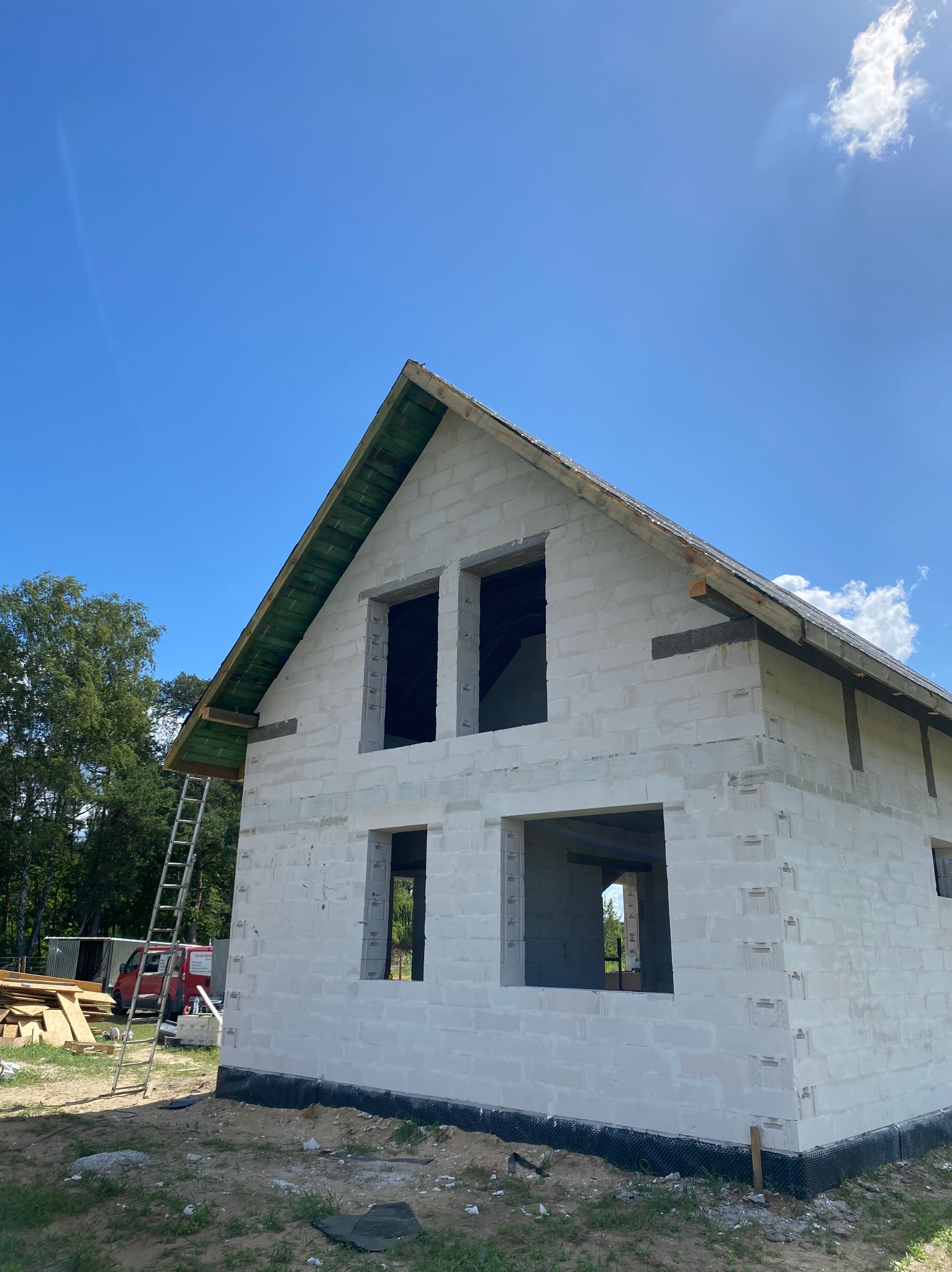 Surowy stan domu z białych bloczków, widoczne otwory okienne i drabina oparta o ścianę, zielone zadaszenie, częściowo wykończony dach w słoneczny dzień.