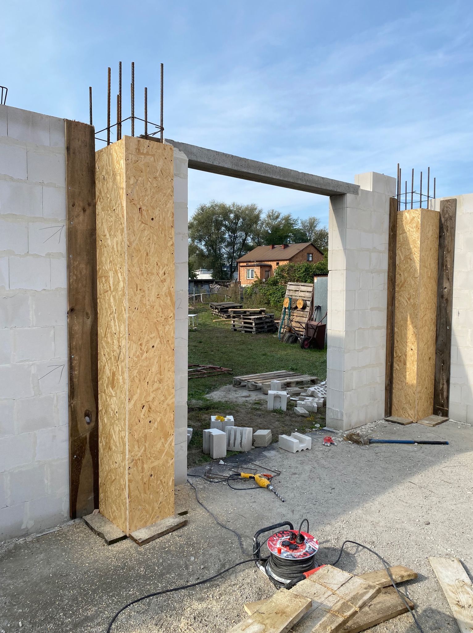 Budowa domu z betonu komórkowego: widok na wznoszone ściany z otworami okiennymi, deskowanie i wystające pręty zbrojeniowe. W tle dom jednorodzinny.