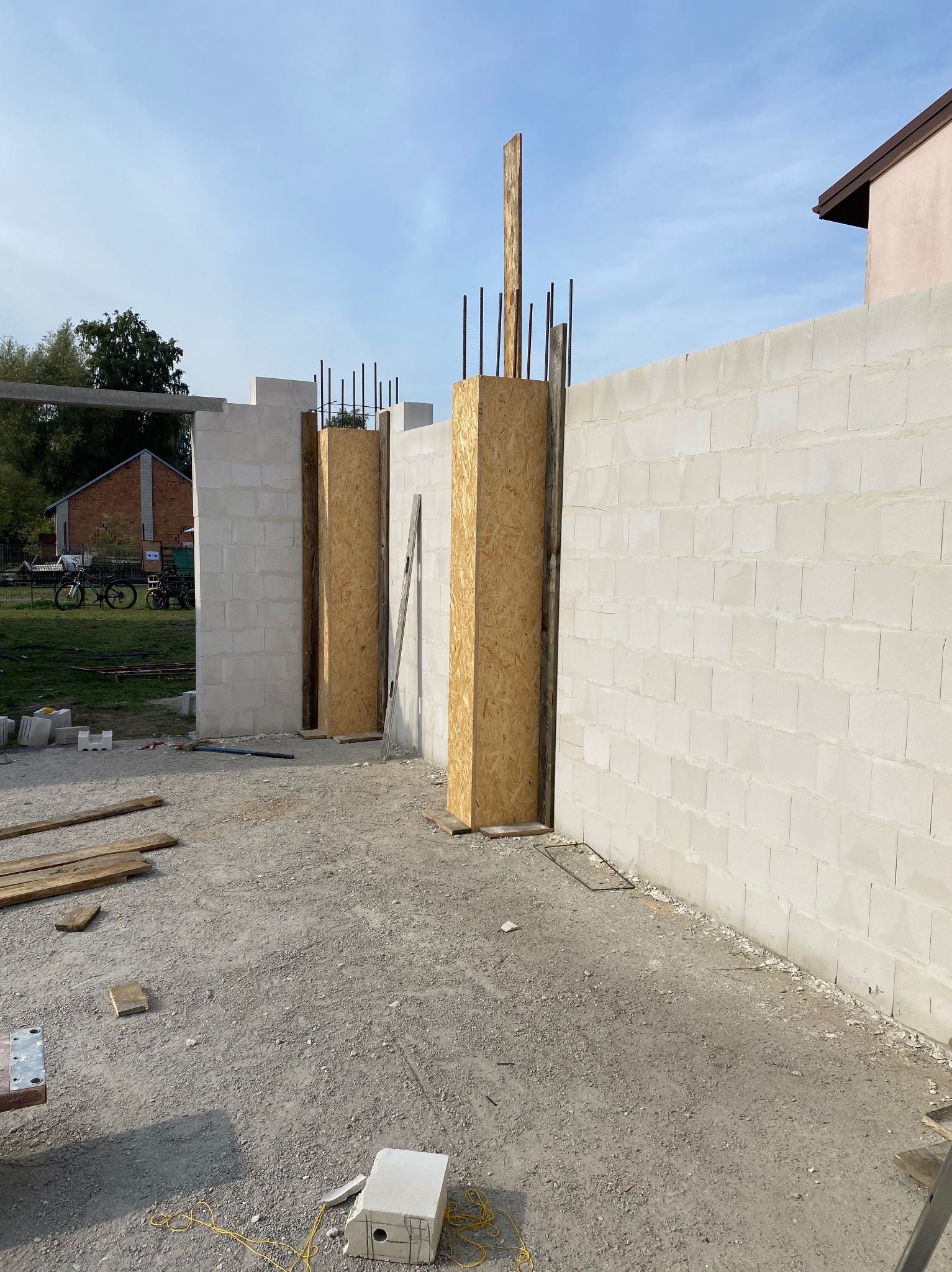 Budowa ściany z bloczków betonowych z drewnianymi szalunkami i wystającymi prętami zbrojeniowymi. Widoczne narzędzia i materiały budowlane na placu budowy.