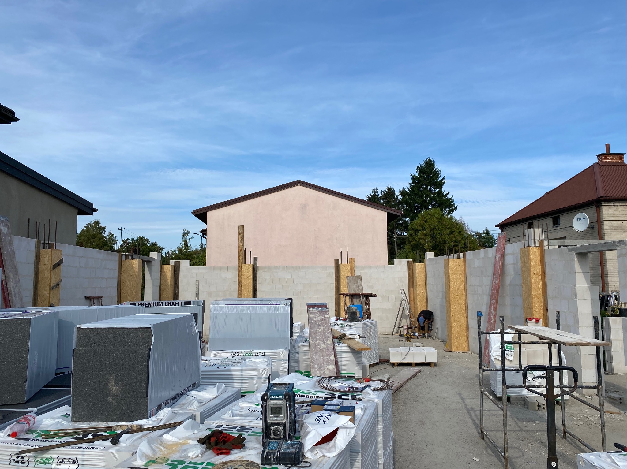 Budowa domu w toku: wzniesione ściany z betonu komórkowego, widoczne materiały izolacyjne Premium Grafit, narzędzia i pracownik w tle. Jasne niebo.