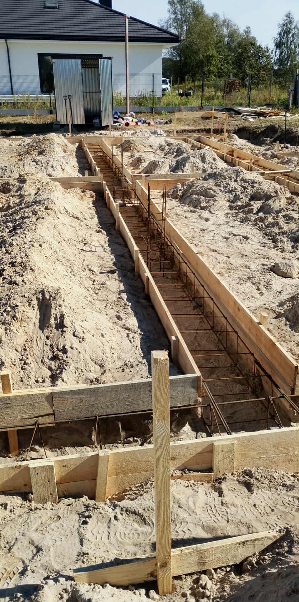 Zbrojenie ławy fundamentowej w drewnianym szalunku na piaszczystym podłożu. Widoczny fragment domu w tle. Prace przygotowawcze do wylania betonu.