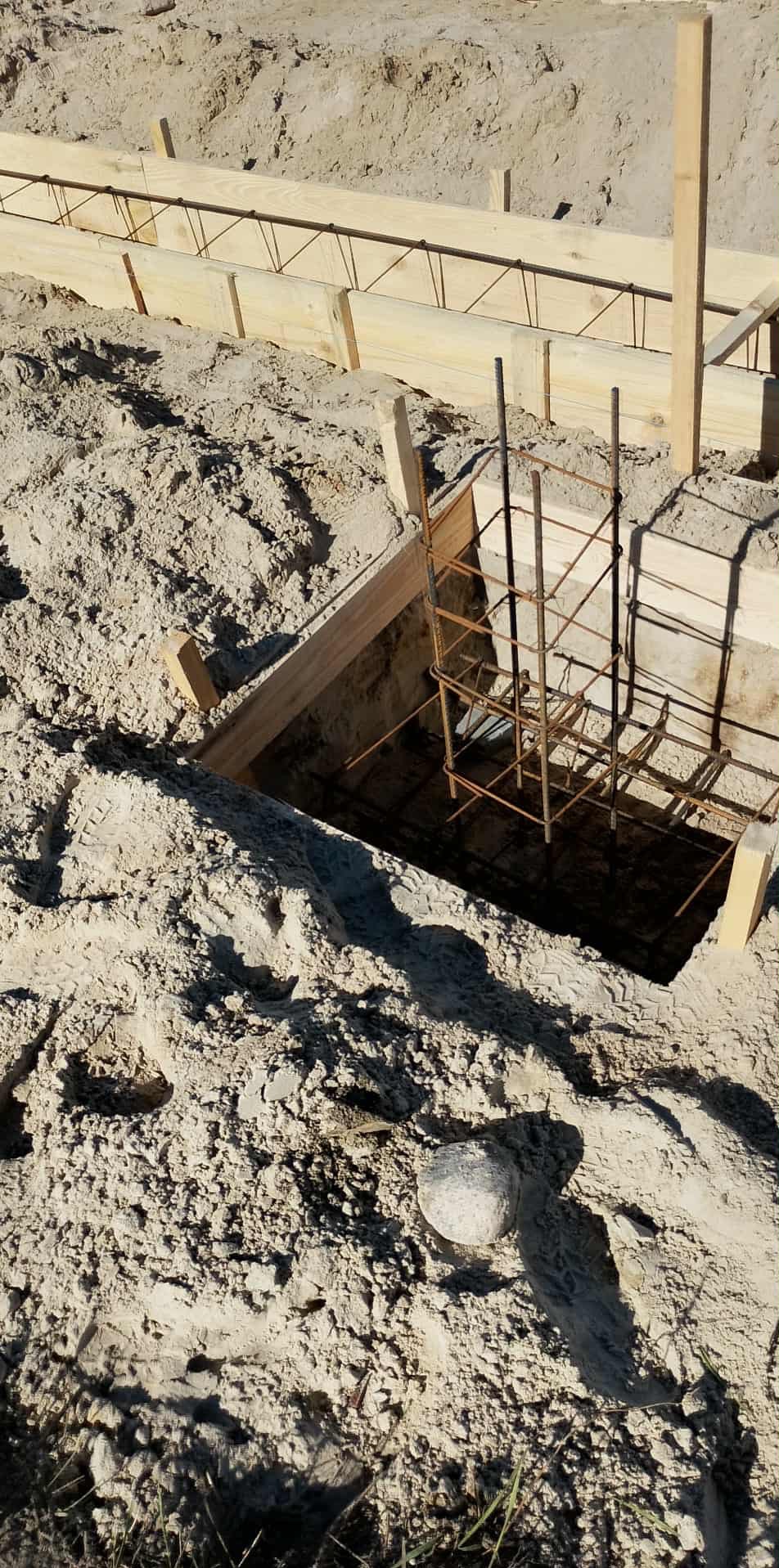 Zbrojenie fundamentu w wykopie z deskowaniem, widoczne pręty stalowe i drewniane szalunki. Ślady opon w piasku. Etap budowy, przygotowanie do zalewania betonem.