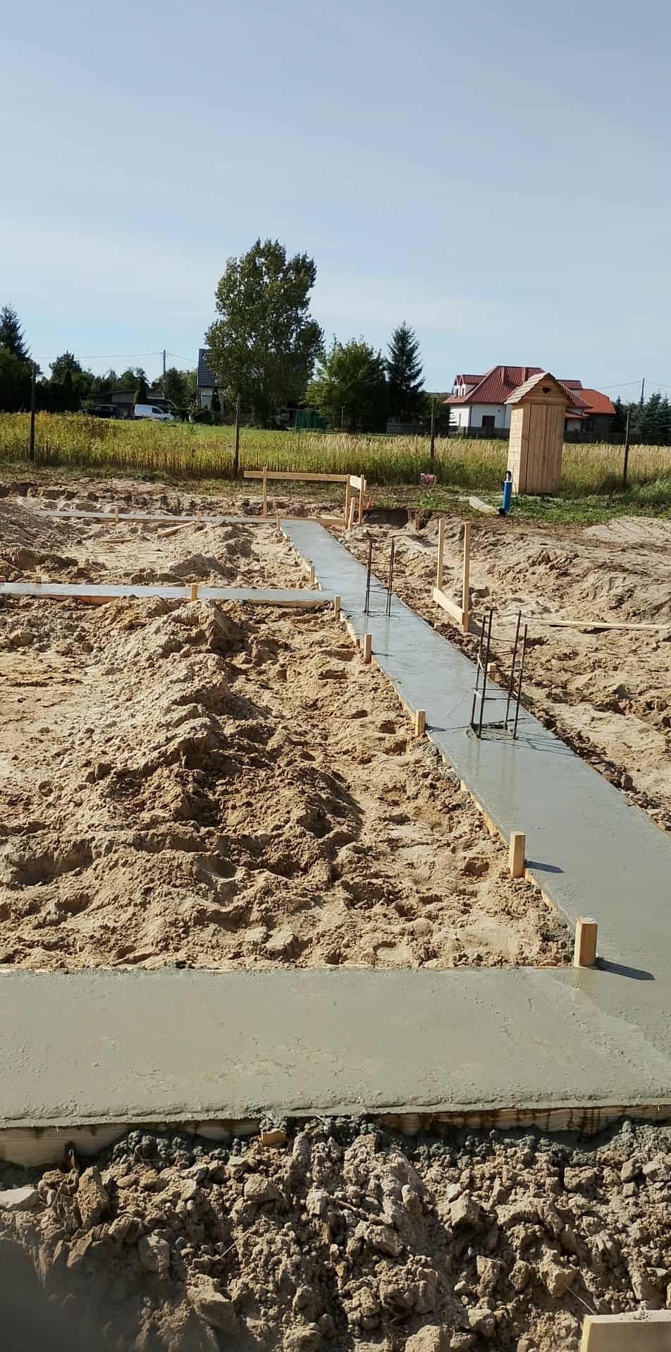 Wylane fundamenty pod budynek, widoczne zbrojenia i szalunki. Tło: pole, drzewa, zabudowania. Ujęcie z poziomu gruntu, słoneczny dzień.