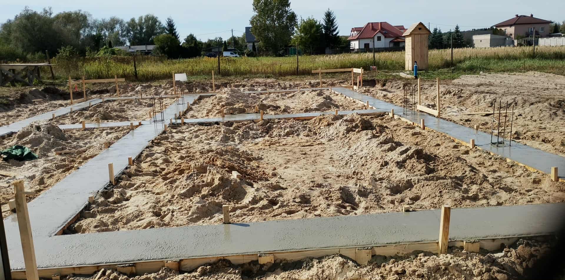 Wylane betonowe ławy fundamentowe na piaszczystym terenie budowy, widoczne drewniane szalunki i wystające pręty zbrojeniowe, dom w tle.