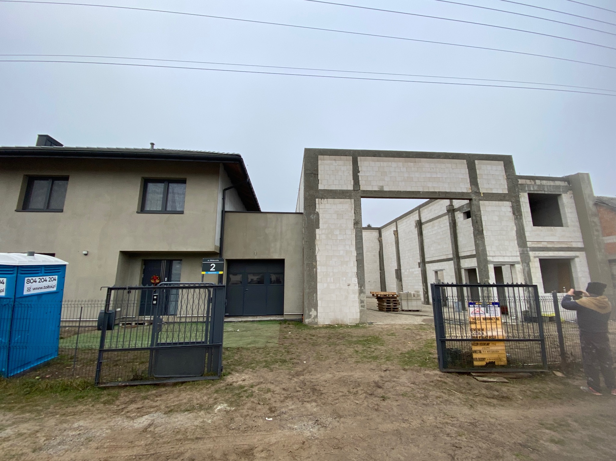 Dom jednorodzinny w trakcie rozbudowy: widoczna część istniejąca z garażem i nowa konstrukcja z betonu komórkowego. Otwarta brama wjazdowa, toaleta przenośna.