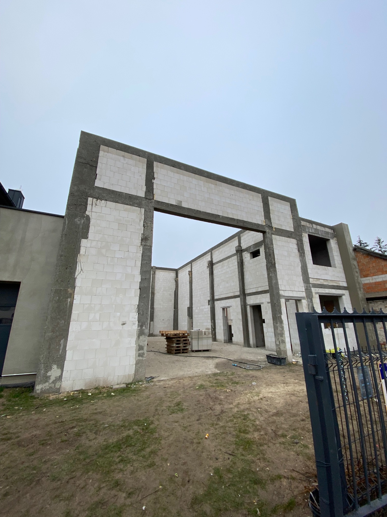 Surowy stan domu z betonu komórkowego w trakcie budowy. Widoczne ściany, otwory okienne i drzwiowe. Na pierwszym planie fragment ogrodzenia.