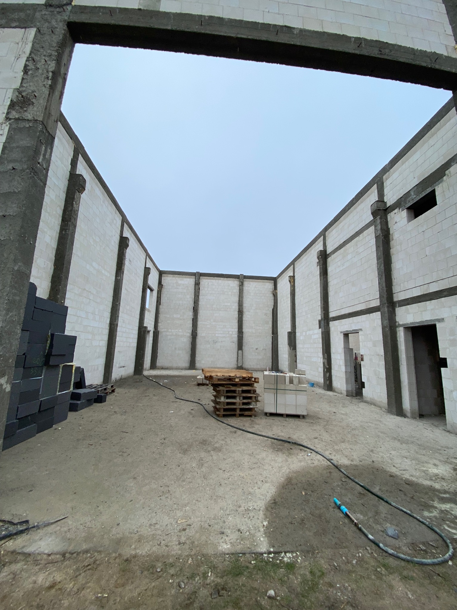 Wnętrze surowego budynku z betonu komórkowego w Lucynowie Dużym, widok z dołu. Widoczne palety z materiałami budowlanymi i wąż. Stan surowy otwarty.
