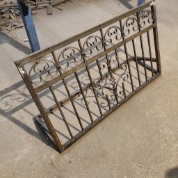 KUNTSTSCHMIEDE BEDNARSKI PIOTR - Fragment metalowej konstrukcji z kutymi zdobieniami, leżący na betonowej podłodze warsztatu, widoczne ślady spawania i obróbki metalu w tle.