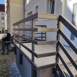 KUNTSTSCHMIEDE BEDNARSKI PIOTR - Nowoczesna, metalowa balustrada schodowa w kolorze grafitowym, zamontowana przed budynkiem, z widocznym fragmentem chodnika z kostki brukowej i sylwetką mężczyzny w tle.