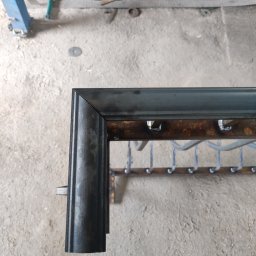 KUNTSTSCHMIEDE BEDNARSKI PIOTR - Fragment metalowej konstrukcji z przyspawanymi elementami, widoczne ślady korozji i spawania, surowy stan wykończenia, warsztatowe otoczenie.