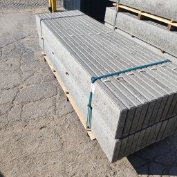 KUNTSTSCHMIEDE BEDNARSKI PIOTR - Betonowe elementy ogrodzeniowe ułożone na palecie, zabezpieczone taśmą, z widocznymi panelami ogrodzeniowymi w tle.