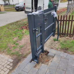 KUNTSTSCHMIEDE BEDNARSKI PIOTR - Szara, metalowa brama automatyczna w trakcie montażu na posesji, widoczny otwarty samochód dostawczy z pracownikiem w tle, kostka brukowa częściowo usunięta.
