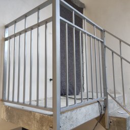 KUNTSTSCHMIEDE BEDNARSKI PIOTR - Szara metalowa balustrada schodowa z pionowymi prętami, zamontowana na surowym betonowym podłożu, widok z dołu na fragment schodów w trakcie remontu.