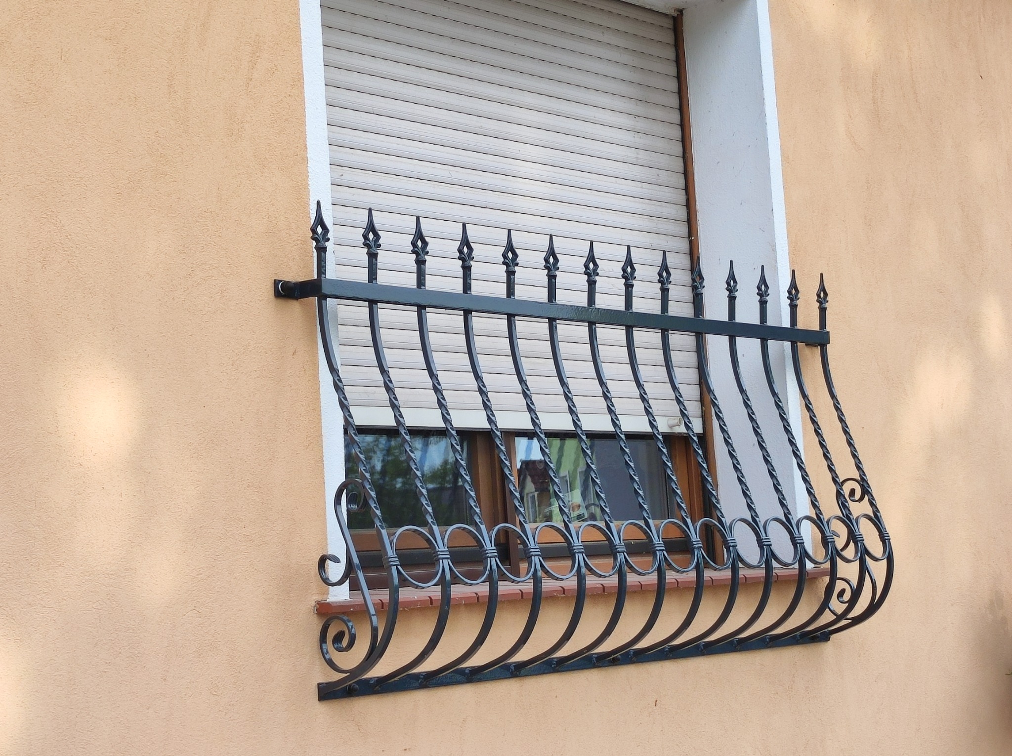Ozdobna, czarna krata okienna z motywem okręgów i grotów na tle beżowej elewacji i zasłoniętego okna.