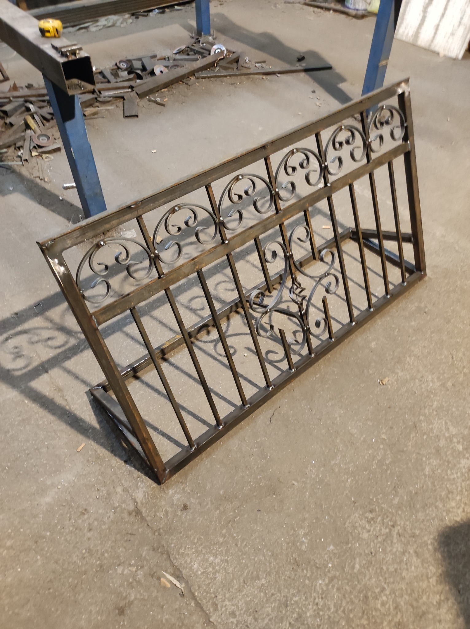 Fragment metalowej konstrukcji z kutymi zdobieniami, leżący na betonowej podłodze warsztatu, widoczne ślady spawania i obróbki metalu w tle.