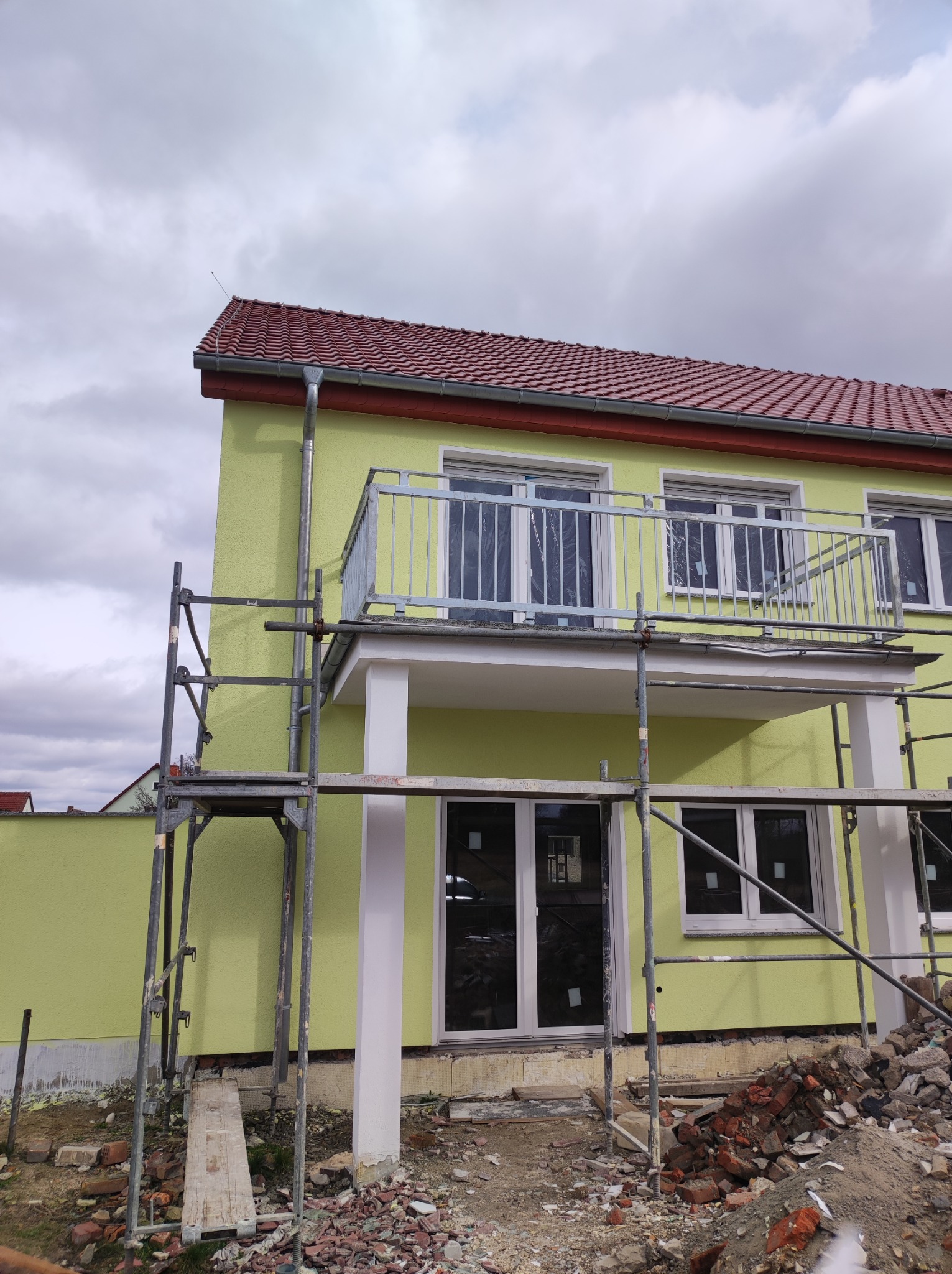 Dom w trakcie budowy lub remontu z żółtą elewacją, metalową balustradą balkonu i rusztowaniem. Widoczne porozrzucane cegły i elementy budowlane.