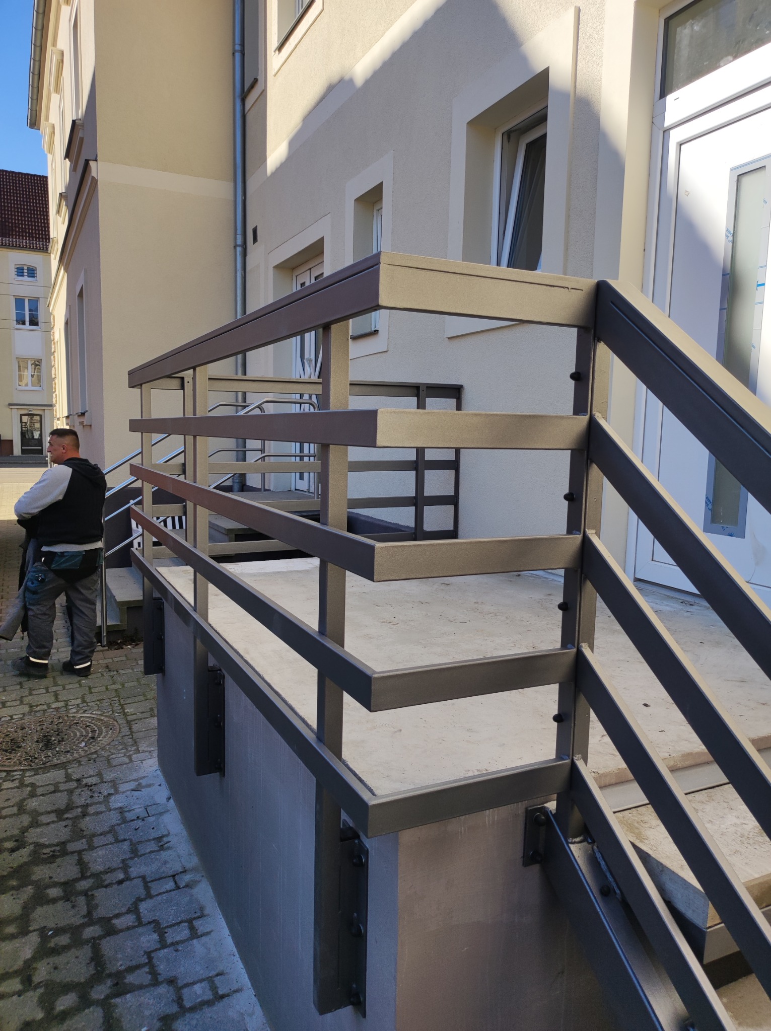 Nowoczesna, metalowa balustrada schodowa w kolorze grafitowym, zamontowana przed budynkiem, z widocznym fragmentem chodnika z kostki brukowej i sylwetką mężczyzny w tle.