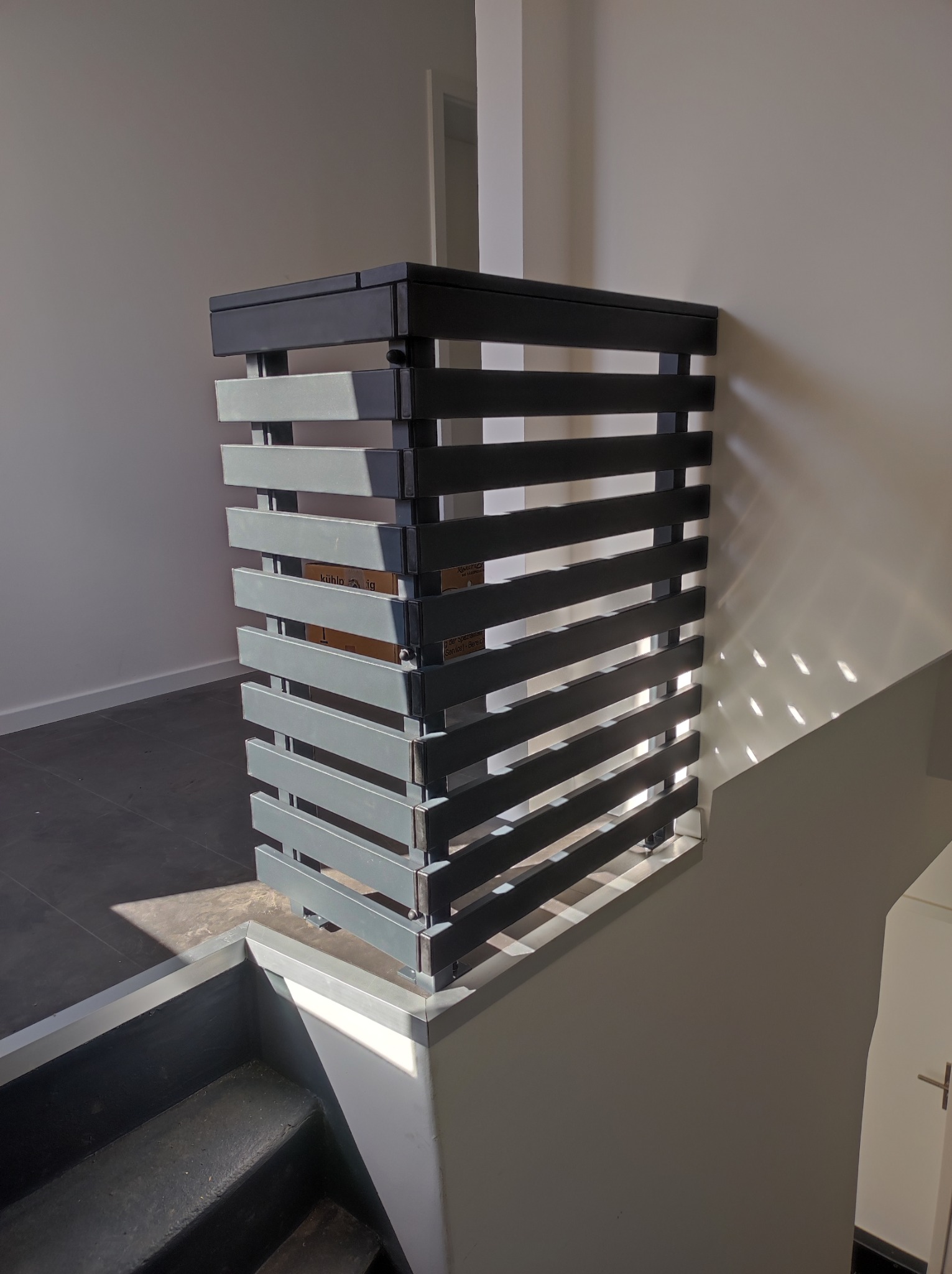 Nowoczesna, pozioma balustrada schodowa z metalowych profili w odcieniach szarości, zamontowana na schodach wewnątrz budynku, z widocznym fragmentem kartonu wewnątrz konstrukcji.