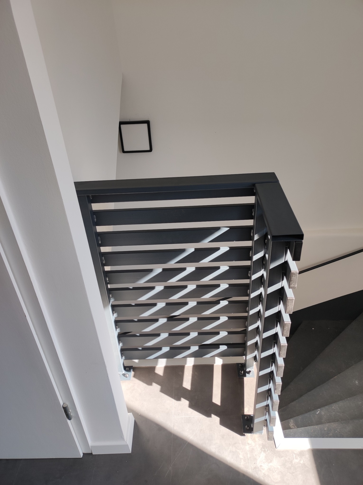 Nowoczesna, metalowa balustrada schodowa z poziomymi panelami, rzucająca geometryczne cienie na szare schody i podłogę, widok z góry.