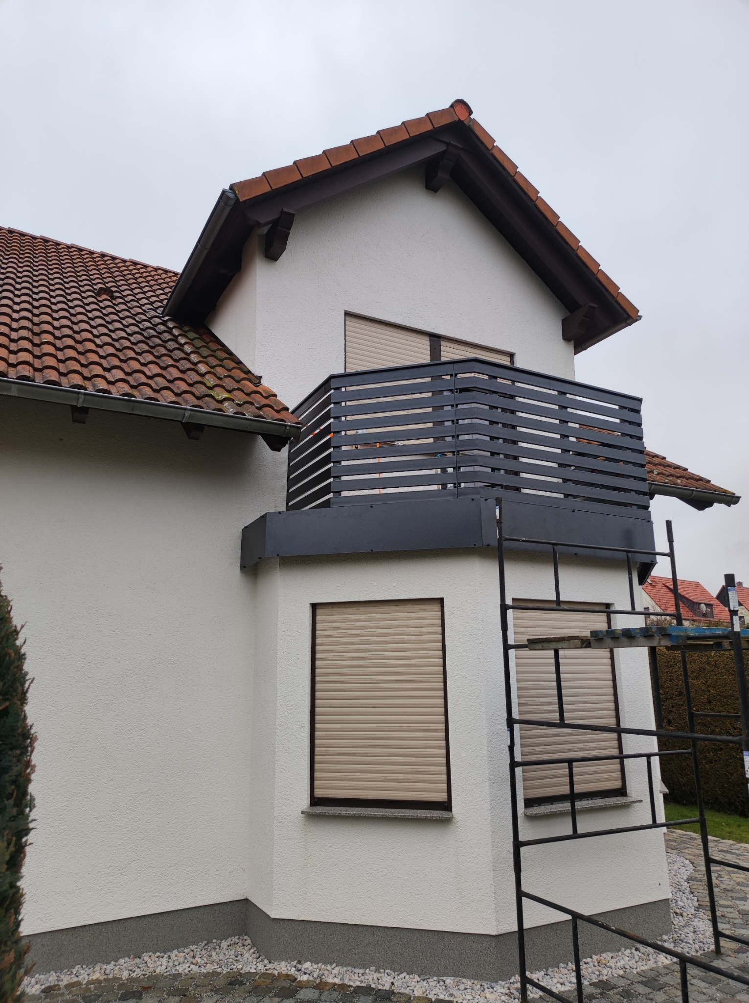 Nowoczesny balkon z poziomą, metalową balustradą w kolorze antracytowym, widok z zewnątrz, z rusztowaniem przylegającym do ściany budynku.