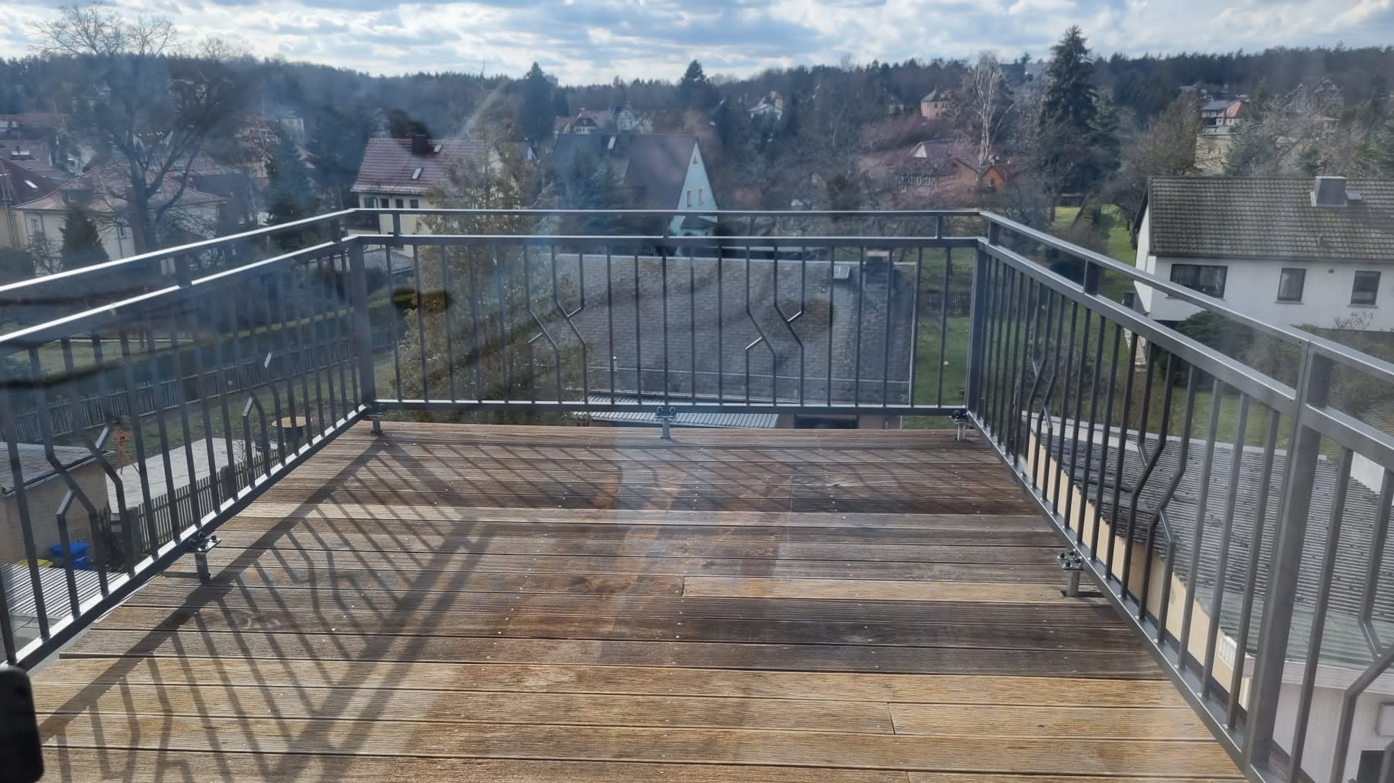 Szare metalowe balustrady balkonu z drewnianą podłogą, widok na zabudowę jednorodzinną i drzewa w tle.