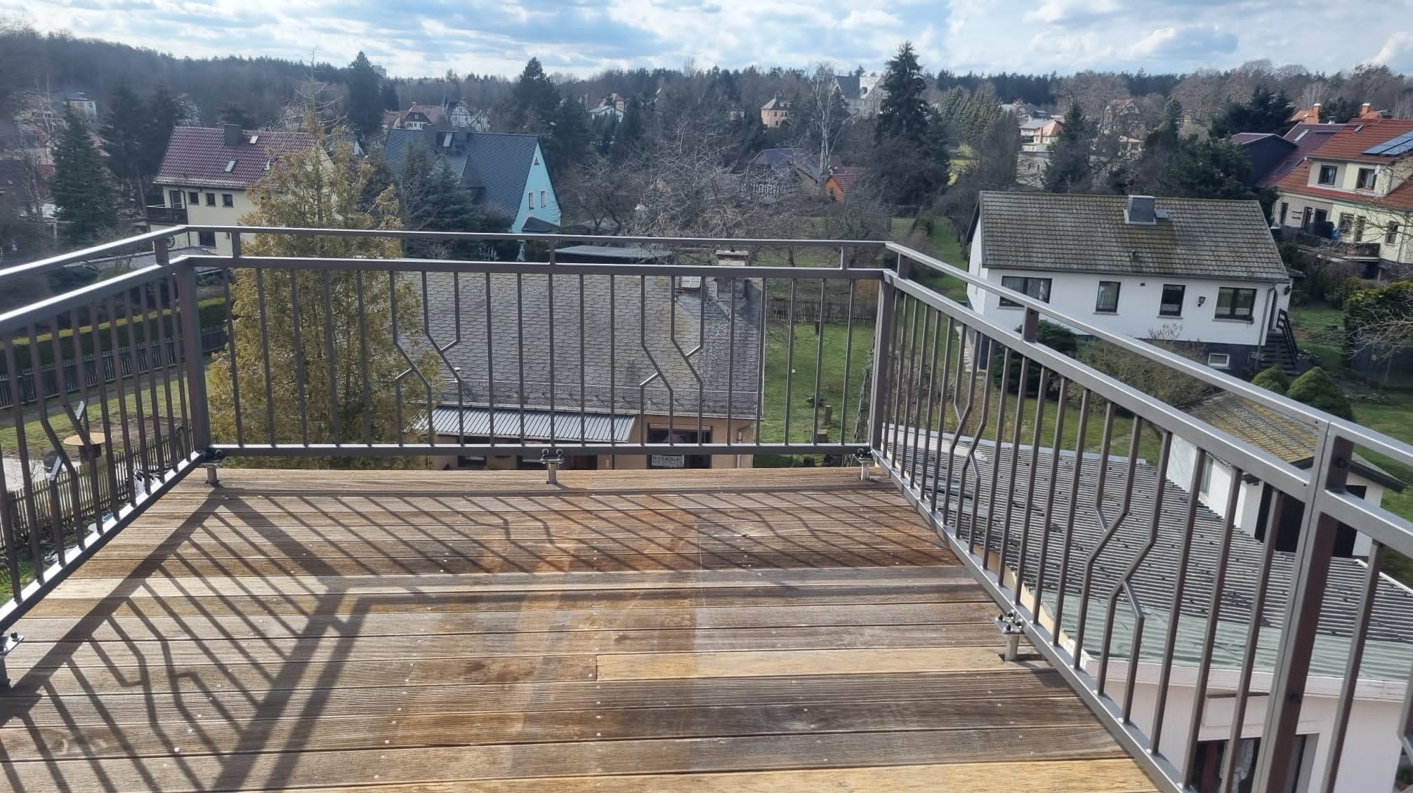 Metalowa balustrada balkonowa z drewnianą podłogą na tle zabudowy jednorodzinnej i drzew.