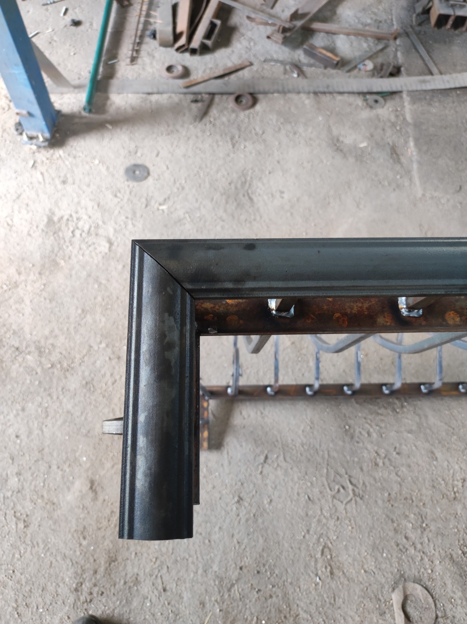 Fragment metalowej konstrukcji z przyspawanymi elementami, widoczne ślady korozji i spawania, surowy stan wykończenia, warsztatowe otoczenie.