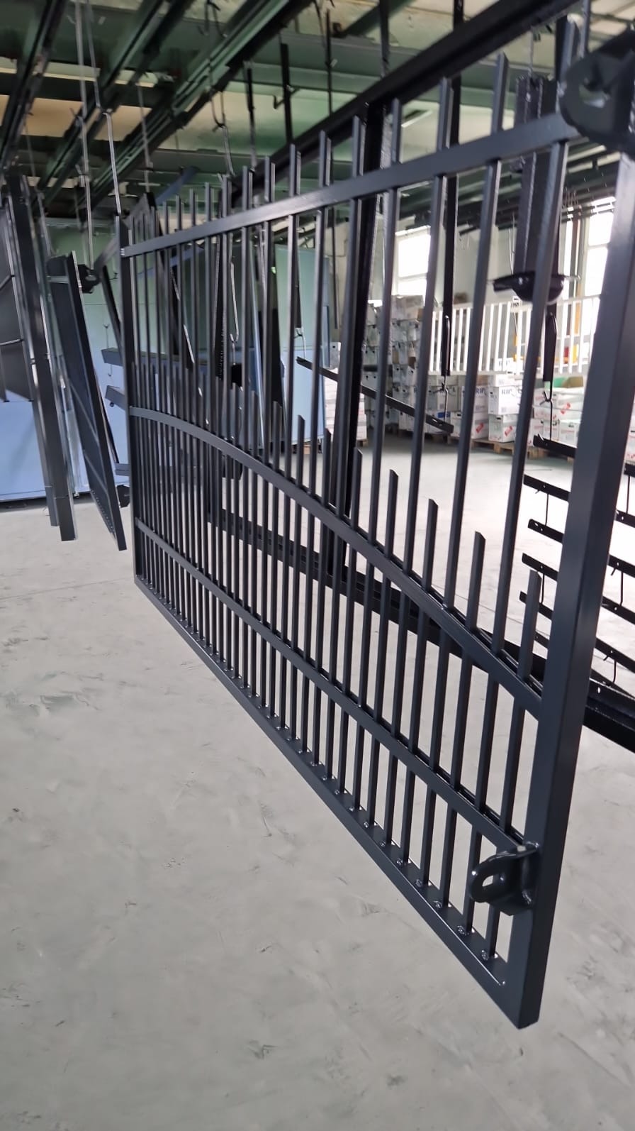 Świeżo pomalowana, metalowa brama w trakcie suszenia na wieszaku w hali produkcyjnej, widoczne elementy konstrukcyjne i uchwyt zamka.