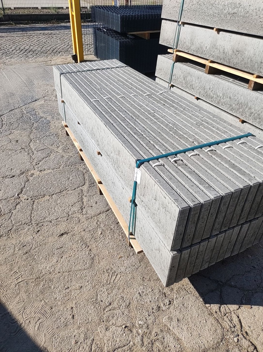 Betonowe elementy ogrodzeniowe ułożone na palecie, zabezpieczone taśmą, z widocznymi panelami ogrodzeniowymi w tle.