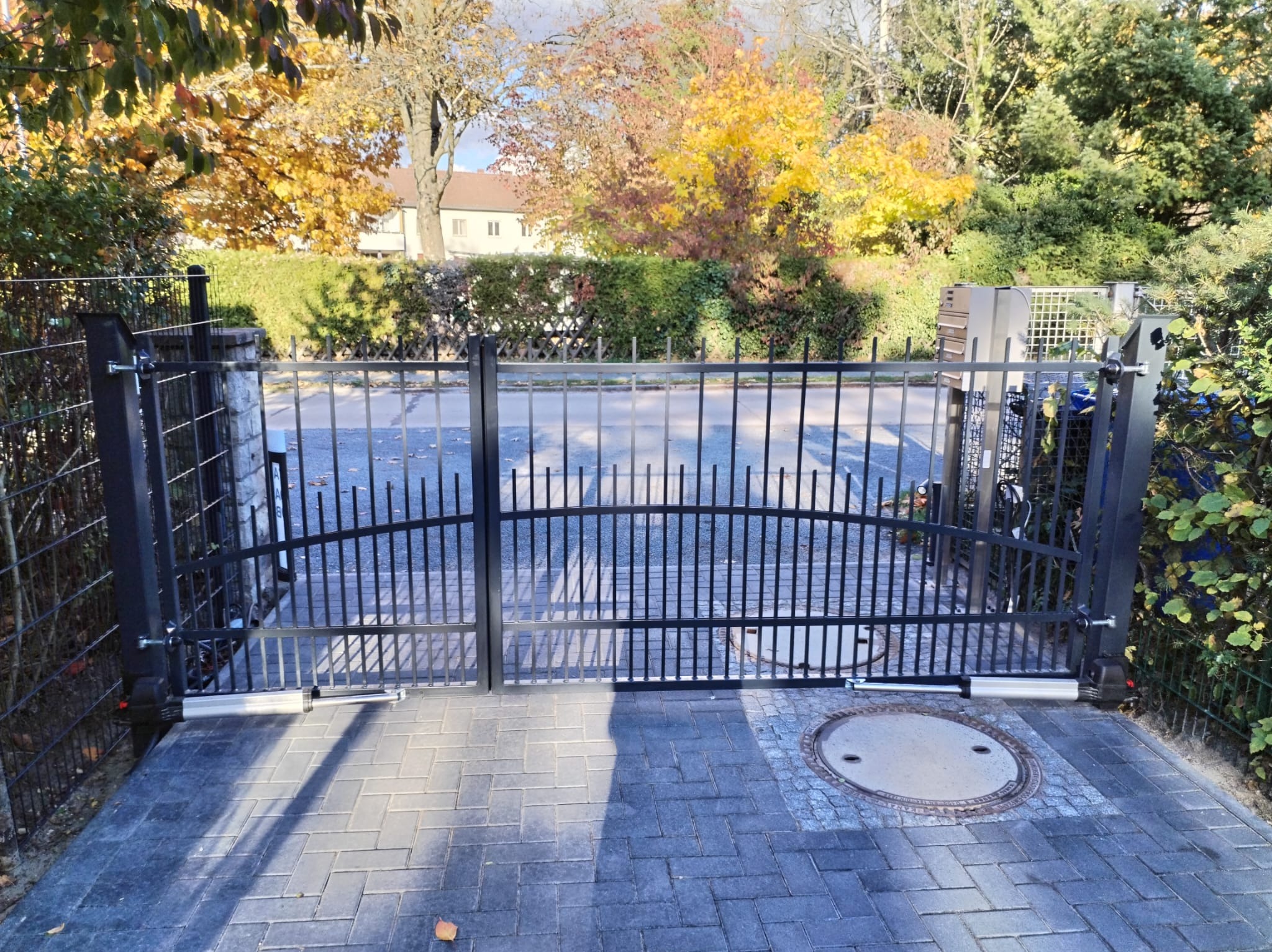 Automatyczna brama wjazdowa z czarnego metalu, widok z zewnątrz, na pierwszym planie kostka brukowa i studzienka kanalizacyjna.