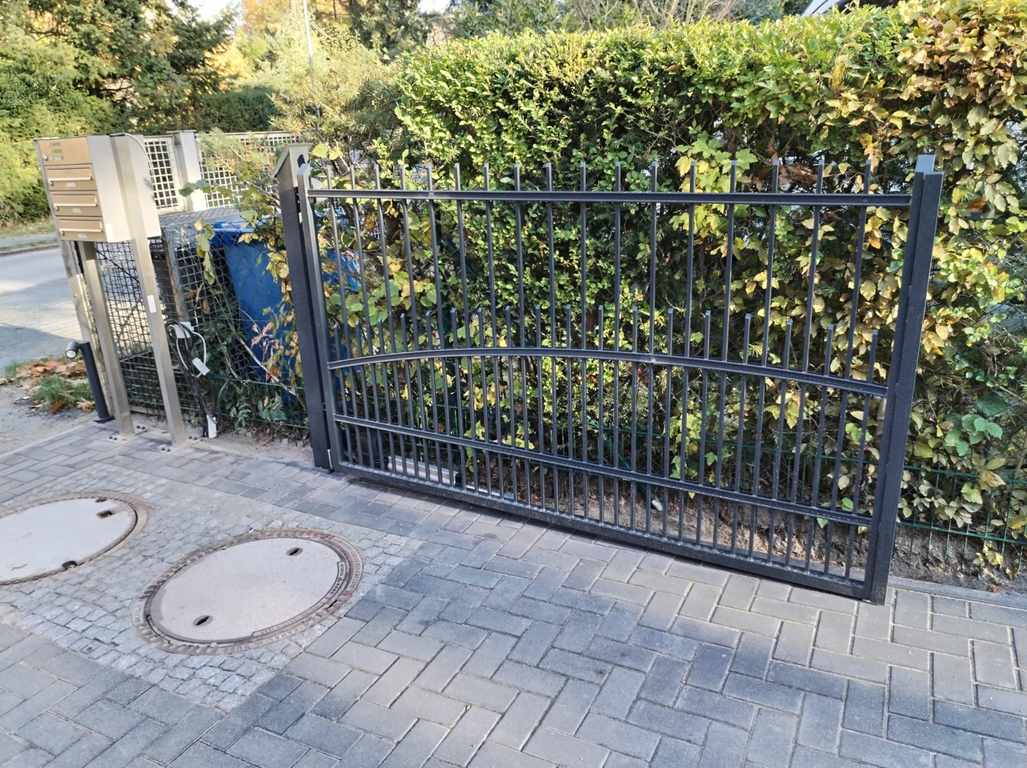 Czarna, metalowa brama wjazdowa z łukowatym wykończeniem górnej części, zamontowana na posesji z kostki brukowej, na tle gęstego, zielonego żywopłotu.