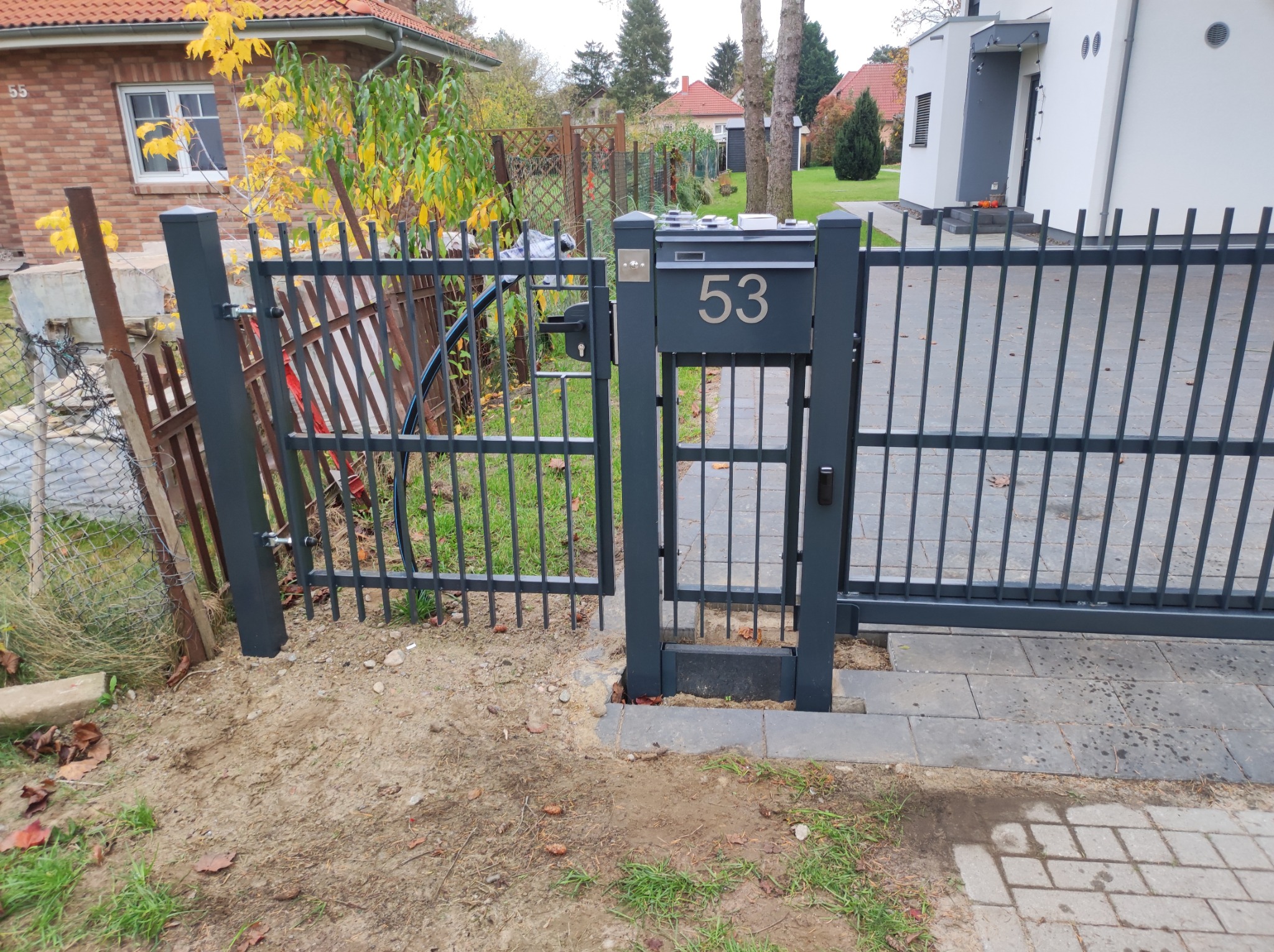 Otwarta, nowoczesna brama wjazdowa z numerem domu 53, po lewej stronie widoczny fragment starego ogrodzenia z siatki.