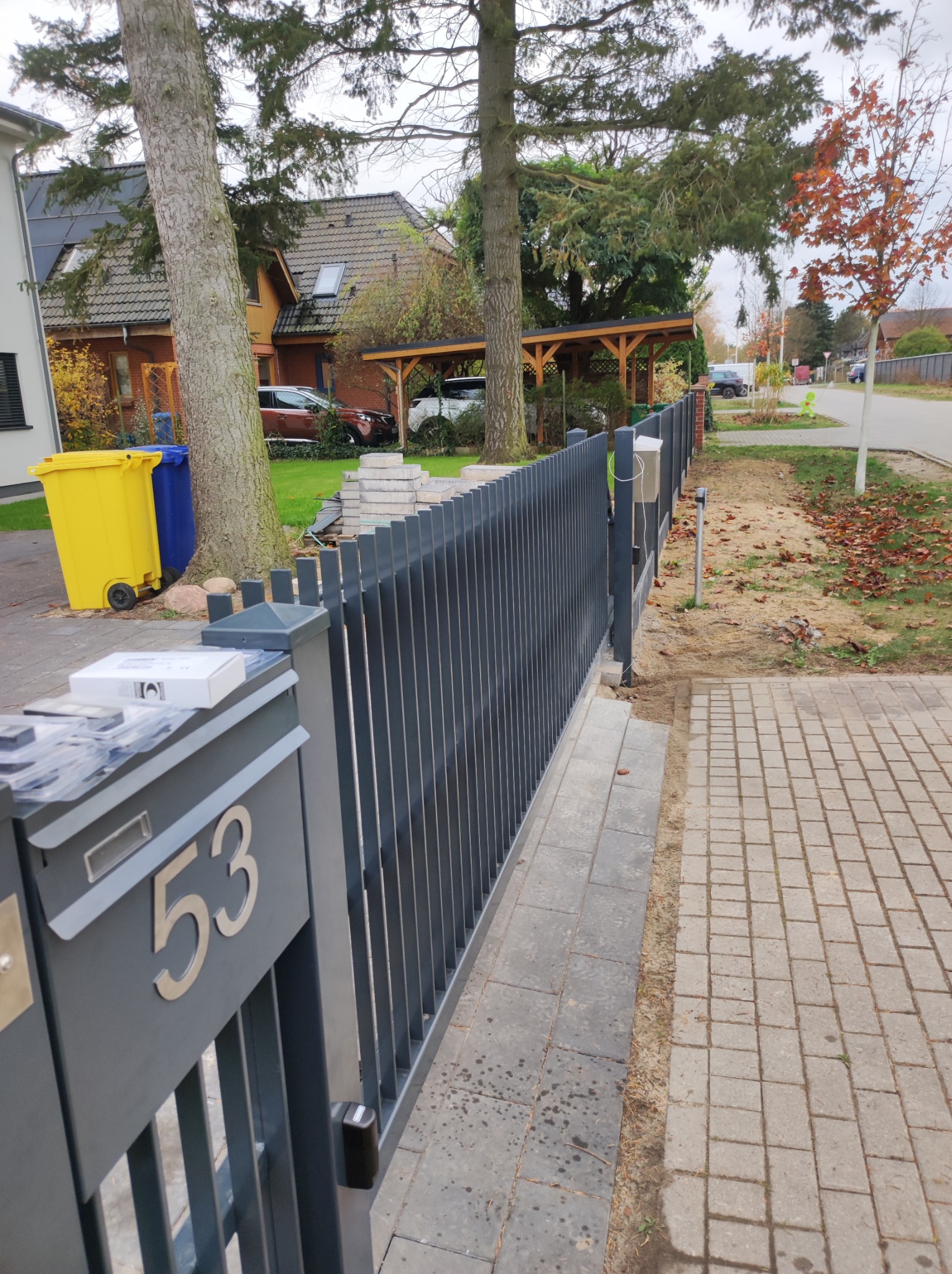 Nowoczesne, pionowe, grafitowe ogrodzenie z metalowymi słupkami, skrzynką pocztową z numerem 53 i automatyką do bramy, widoczne kosze na śmieci i zaparkowane auta w tle.