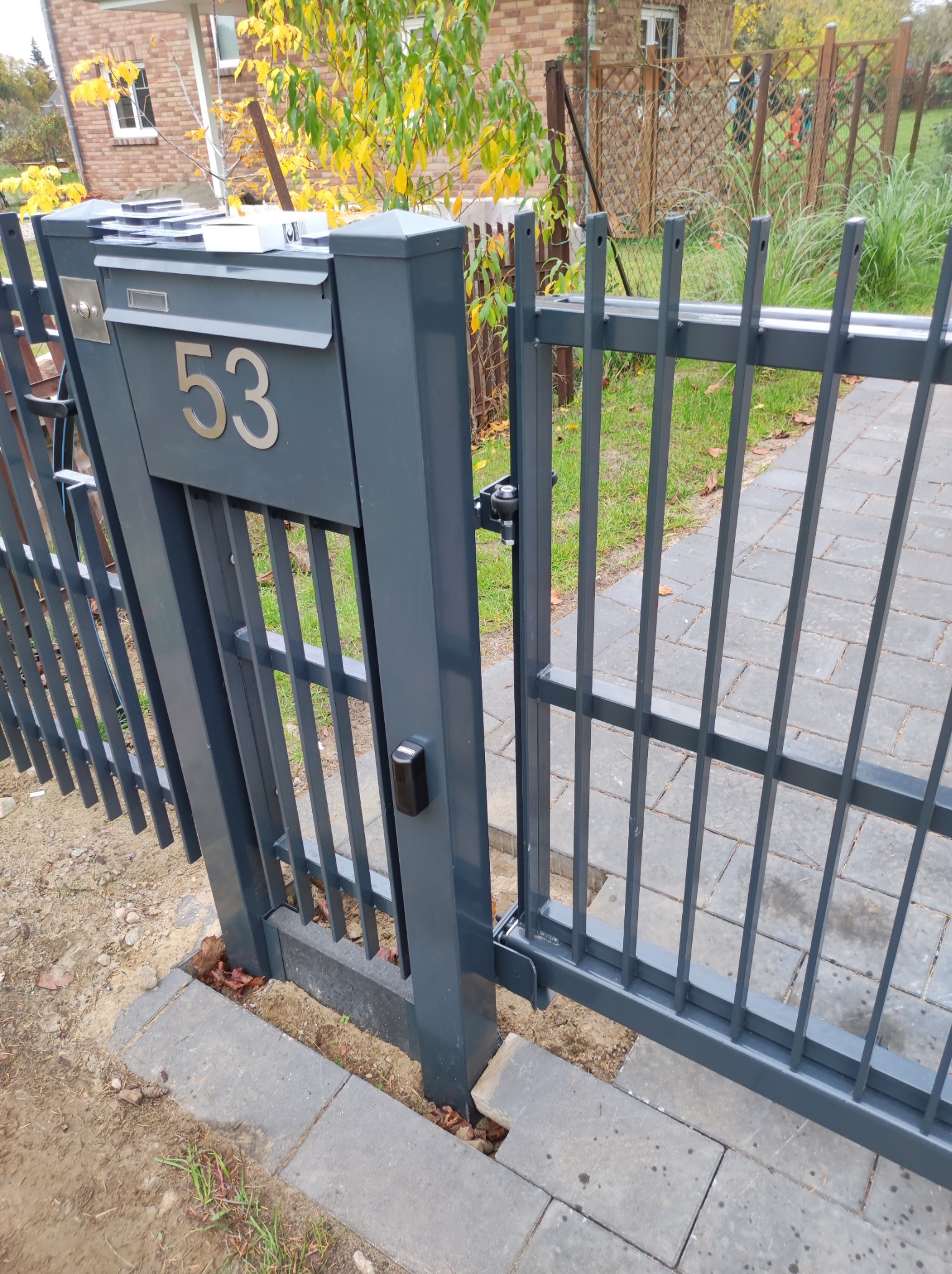 Szara, metalowa brama z numerem domu '53' i skrzynką na listy, widok z bliska na mechanizm otwierania i fragment posesji z kostki brukowej.