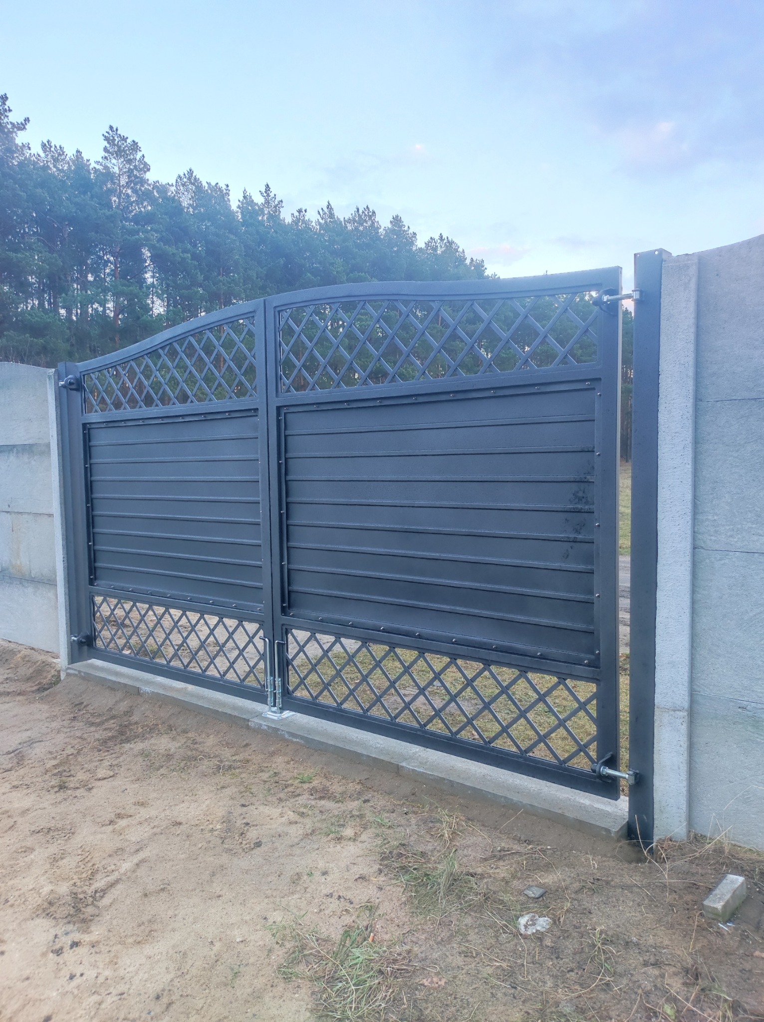 Zamykana, przesuwna brama wjazdowa z metalowymi przęsłami o poziomym układzie i kratownicą, zamontowana między betonowymi słupkami na tle lasu.