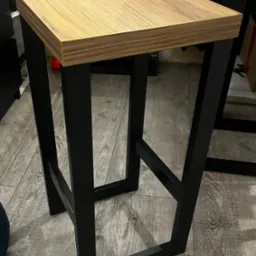 Nowoczesny taboret barowy z blatem z jasnego drewna i czarną, metalową konstrukcją, stojący na podłodze z paneli imitujących drewno.