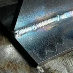 Zbliżenie na świeży spaw dwóch metalowych elementów, widoczne tęczowe przebarwienia na spoinie i powierzchni metalu.