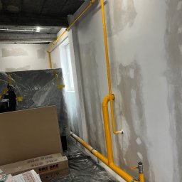 Paweł Goździewicz - Żółta instalacja gazowa poprowadzona wzdłuż ściany w surowym stanie, widoczne niedoskonałości tynku, rury mocowane do ściany za pomocą uchwytów, w tle okno i zakryte folią meble.
