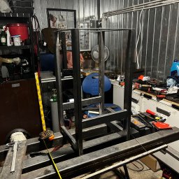 Paweł Goździewicz - Konstrukcja metalowa w trakcie spawania w warsztacie, widoczny spawacz, butla z gazem, miara i narzędzia na stole roboczym.