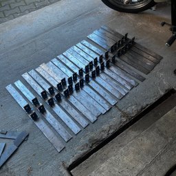 Paweł Goździewicz - Równy rząd metalowych płaskowników z przyspawanymi uchwytami, ułożonych na betonowej podłodze garażu obok motocykla, gotowych do montażu.
