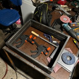 Paweł Goździewicz - Metalowa rama spawana z profili, zabezpieczona zaciskami stolarskimi, widoczna na warsztatowym stole z narzędziami i akcesoriami do obróbki metalu.