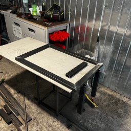 Paweł Goździewicz - Stół warsztatowy z metalowym stelażem w trakcie spawania, widoczne profile stalowe, spawarka kątowa na podłodze, tło z blachy falistej.