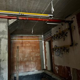 Paweł Goździewicz - Surowy stan pomieszczenia w trakcie instalacji: widoczne rury gazowe w kolorze żółtym i czerwonym, mocowane do sufitu i ściany, z licznymi zaworami i manometrem, ściany częściowo wykończone...