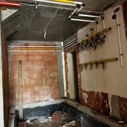 Paweł Goździewicz - Wnętrze w trakcie remontu z widocznymi instalacjami gazowymi i wodnymi, ściany częściowo w surowym stanie, widoczne rury w kolorach żółtym, białym i czerwonym, zamocowane do ściany i sufitu...