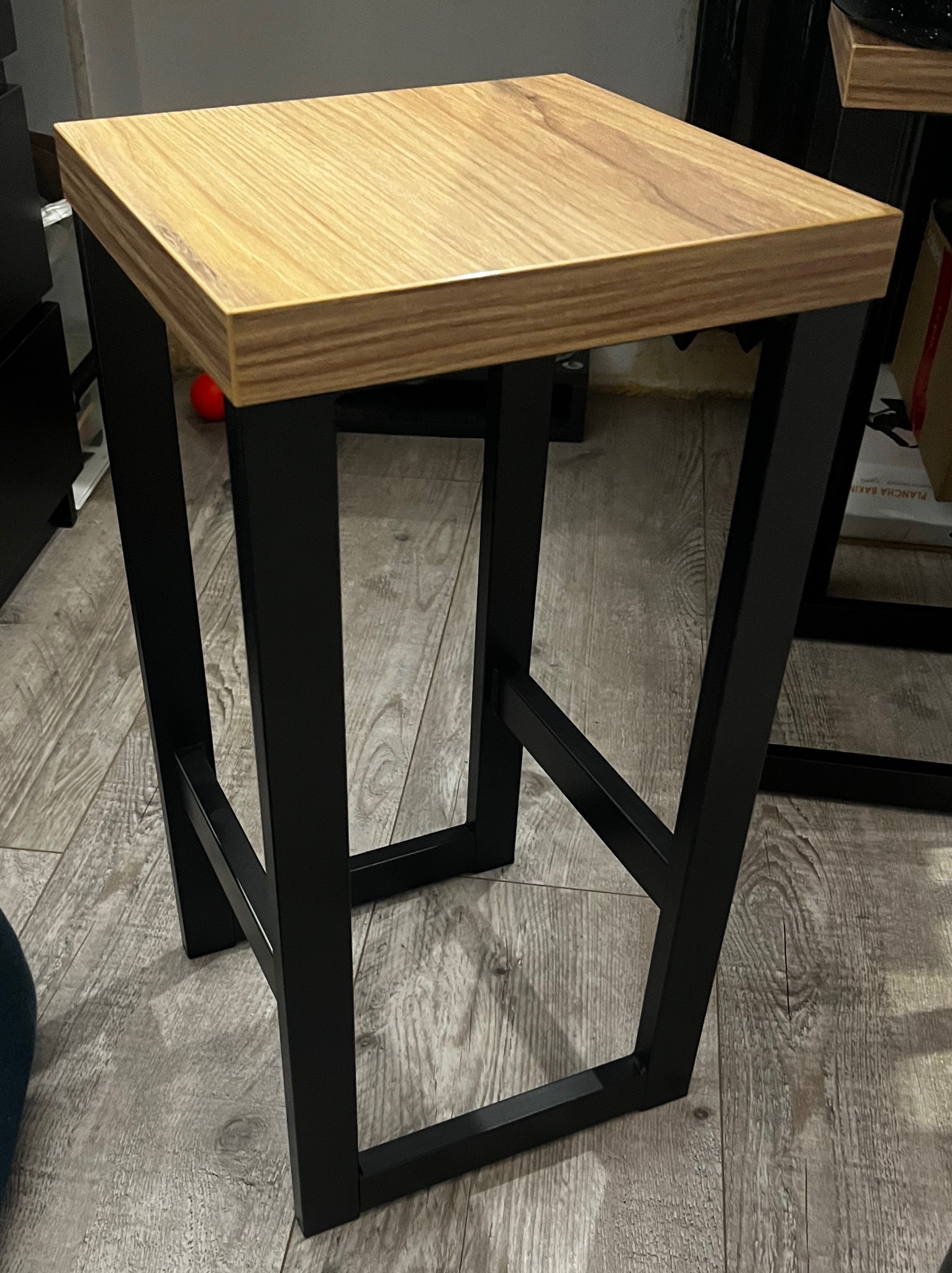 Nowoczesny taboret barowy z blatem z jasnego drewna i czarną, metalową konstrukcją, stojący na podłodze z paneli imitujących drewno.